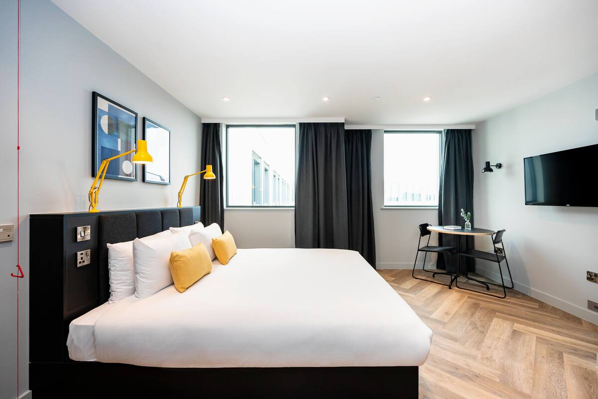 A modern hotel room with a large bed, two bedside lamps, a flat-screen TV, a small table with chairs, and large windows with curtains.