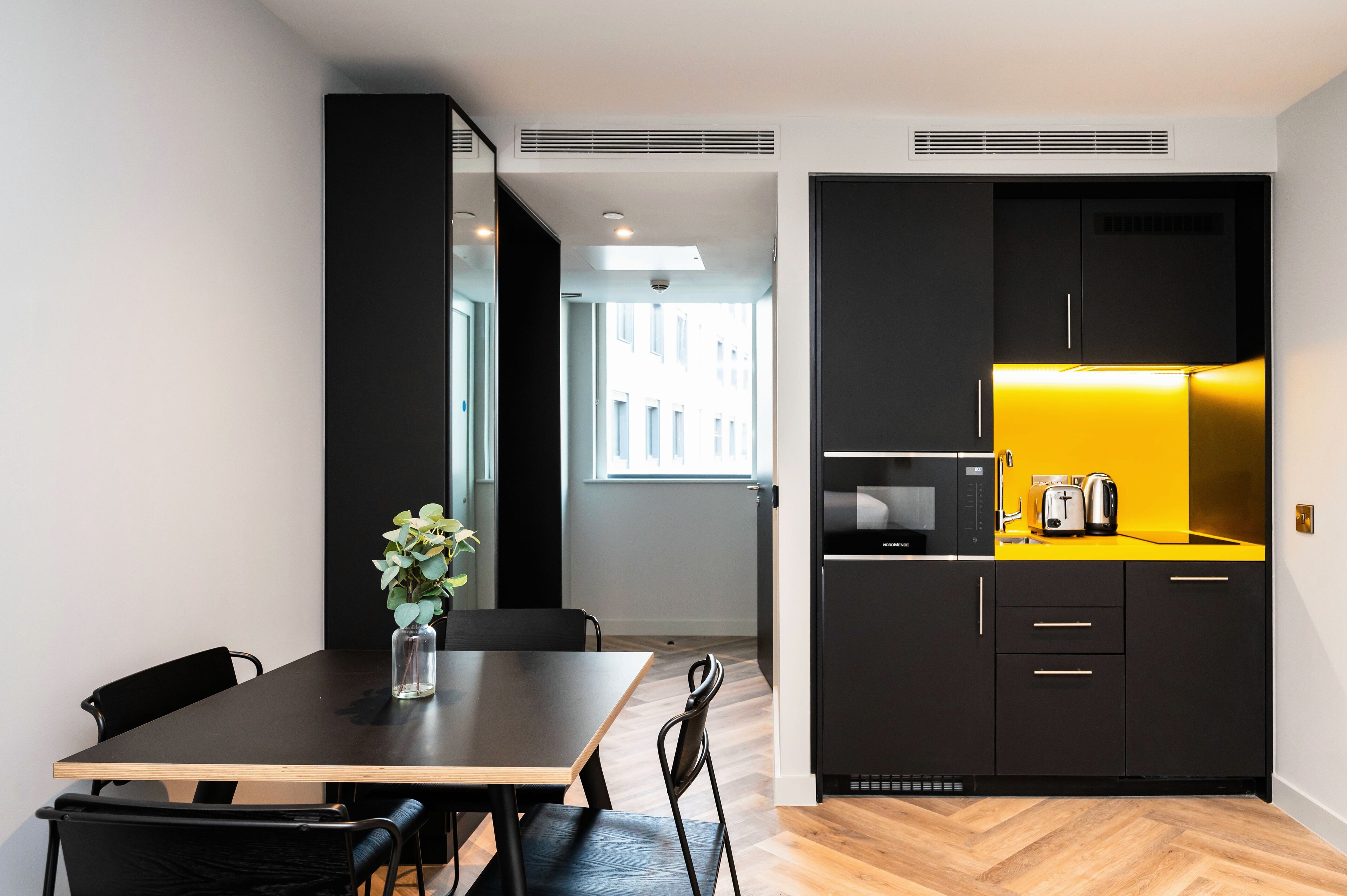 A modern kitchen with black cabinets and a yellow countertop, a dining area with a wooden table and black chairs, and a view of the city through a window.