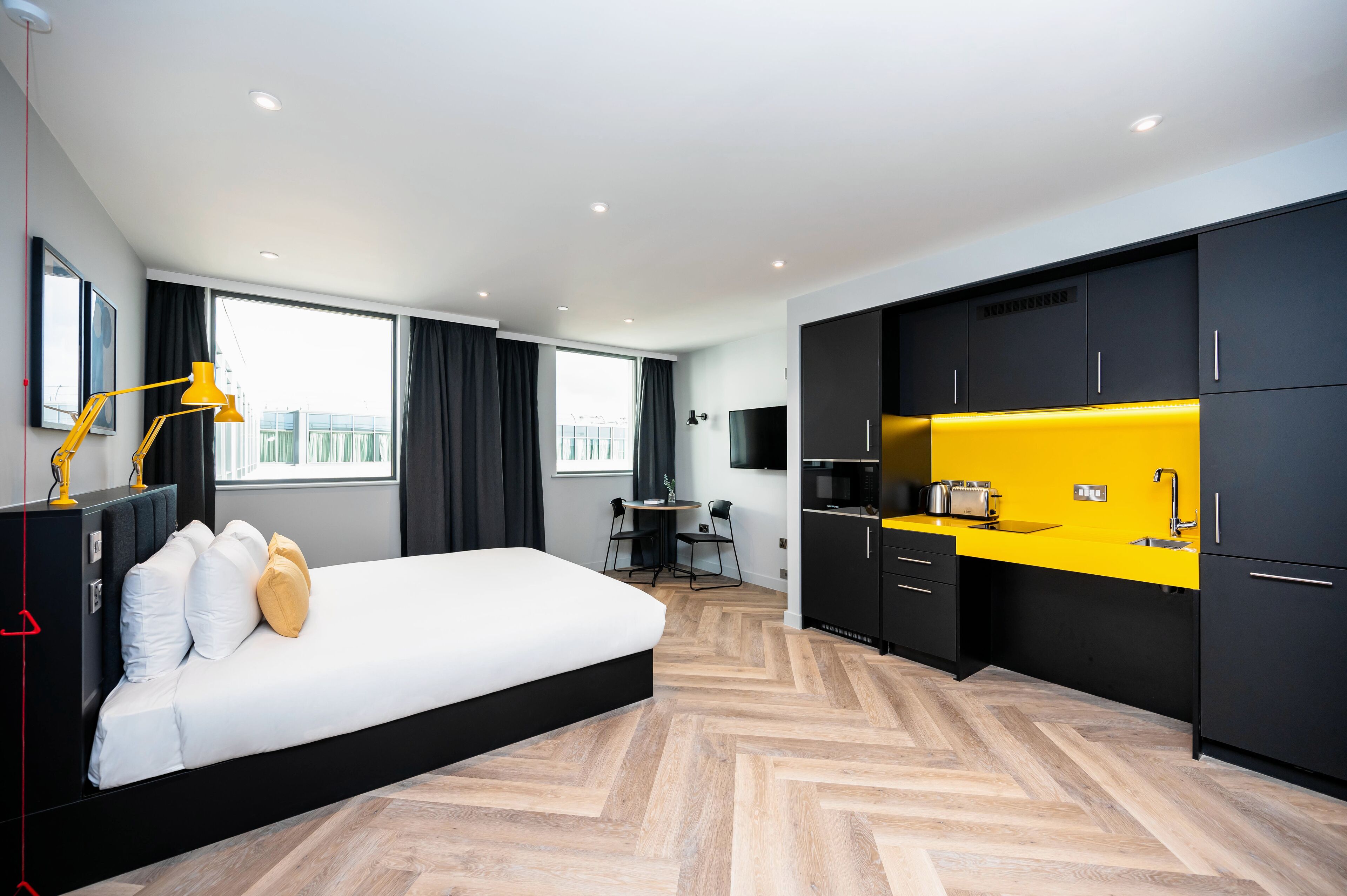 A modern hotel room with a bed, a desk, a chair, a small kitchen area, and a large window.