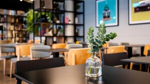 Un espacio interior moderno con mesas y sillas, un jarrón de cristal con plantas verdes y una estantería al fondo.