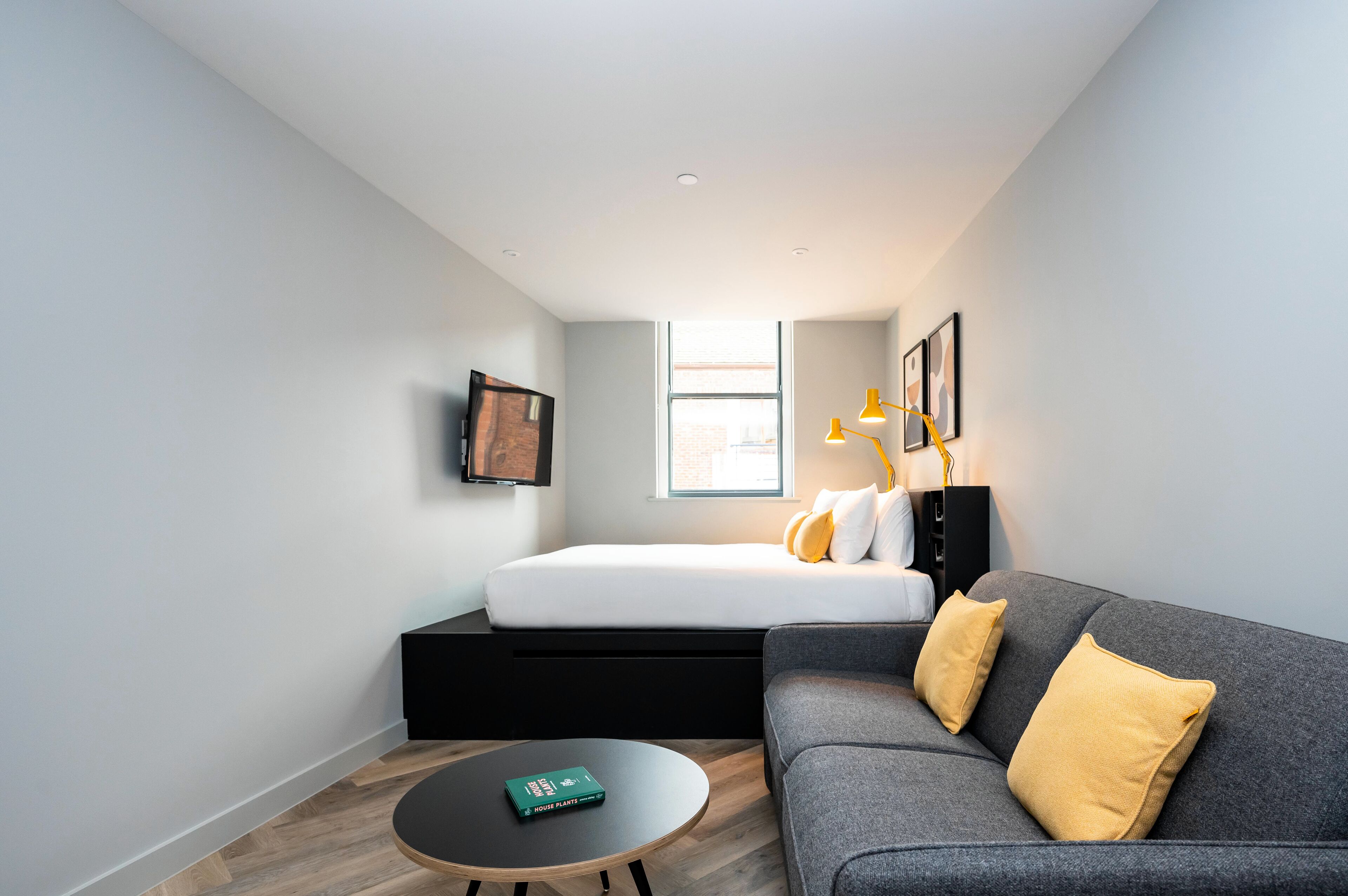 Une chambre moderne et compacte, dotée d’un lit, d’un canapé gris, d’une petite table ronde et d’une fenêtre laissant entrer la lumière naturelle.