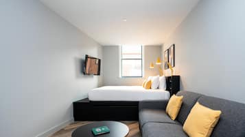 Une chambre moderne et compacte, dotée d’un lit, d’un canapé gris, d’une petite table ronde et d’une fenêtre laissant entrer la lumière naturelle.