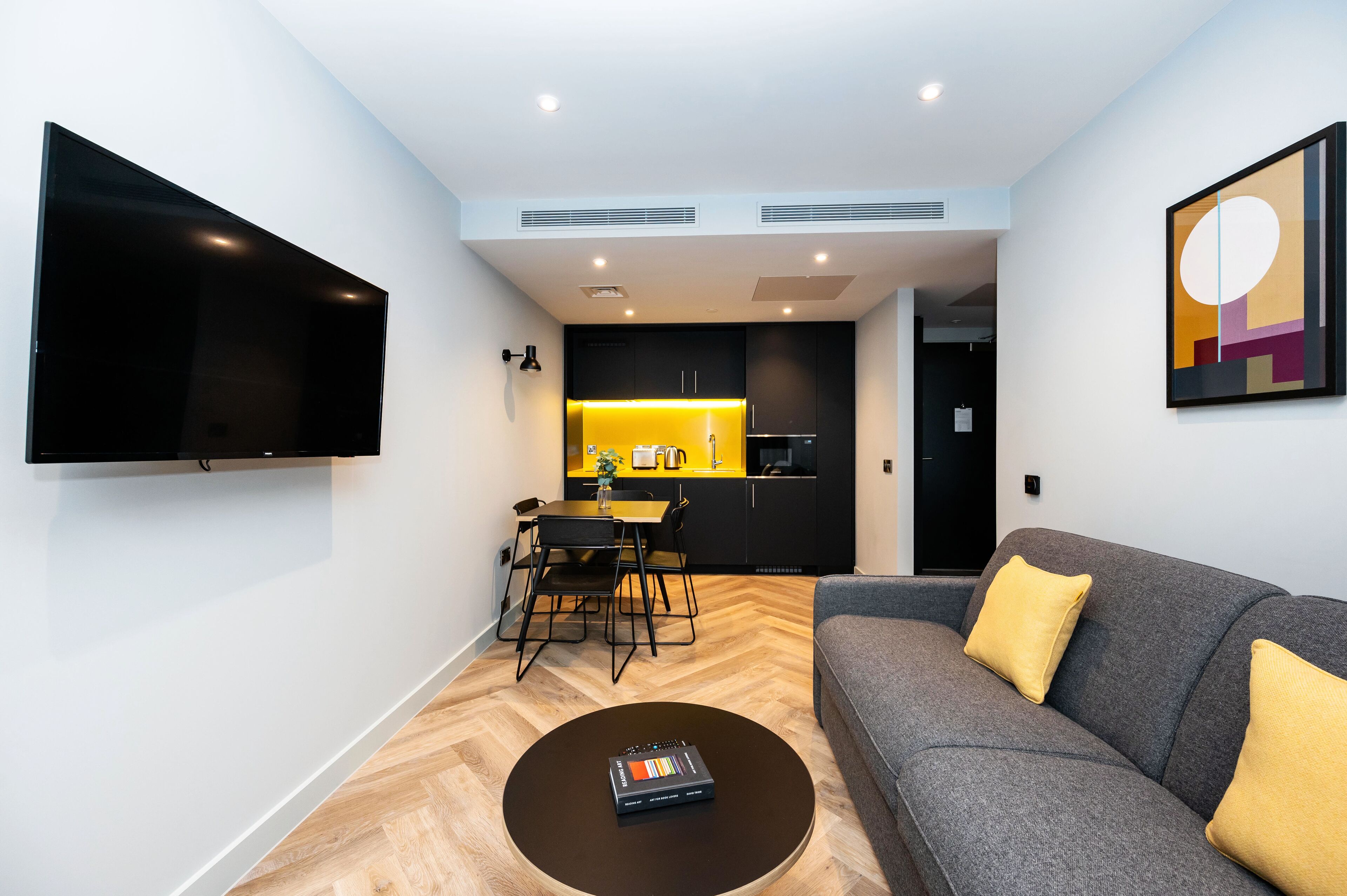 A modern living room with a flat-screen TV, a sofa with yellow cushions, a coffee table, and a dining area with a yellow-lit kitchen in the background.