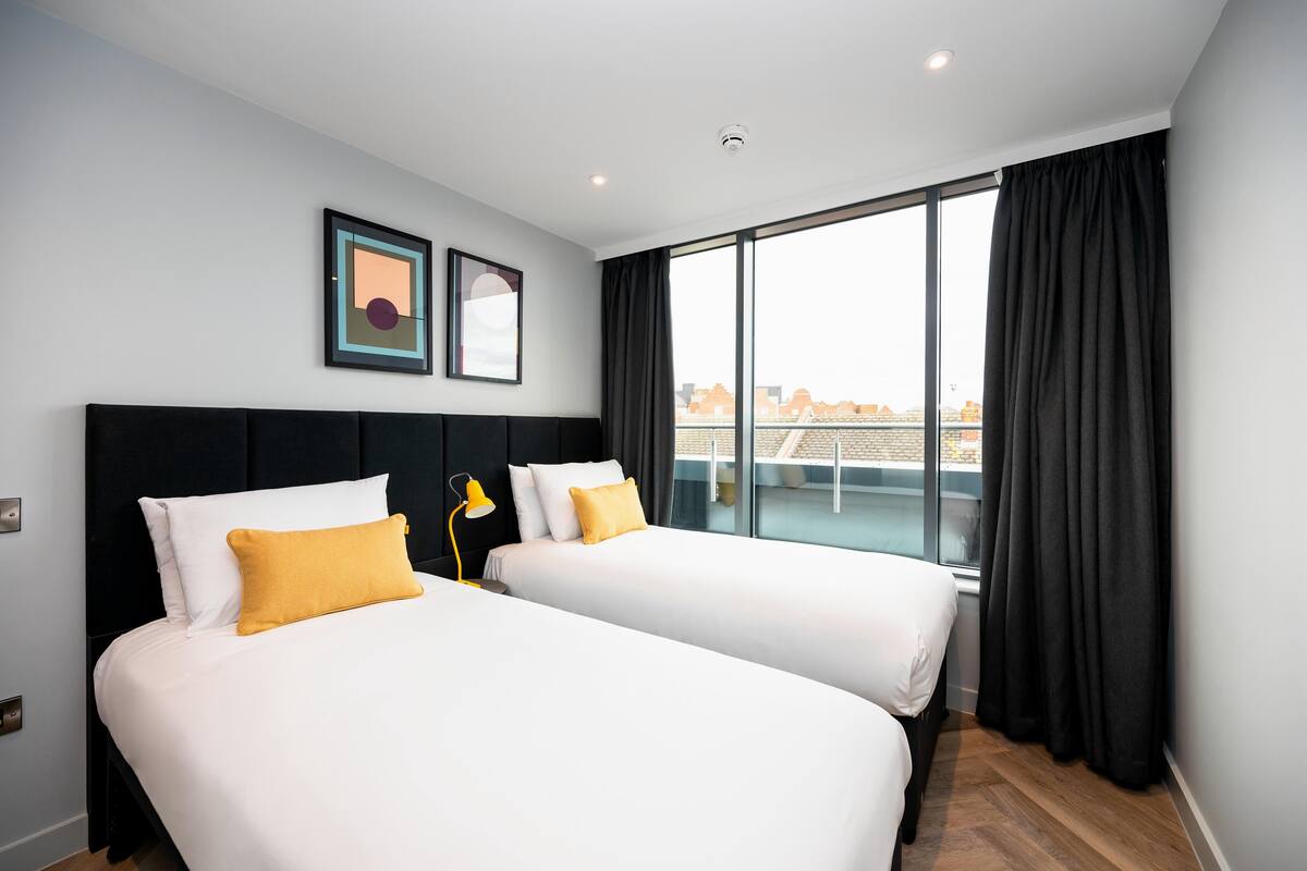 A hotel room with two beds, a large window with curtains, a city view, and a framed picture on the wall.