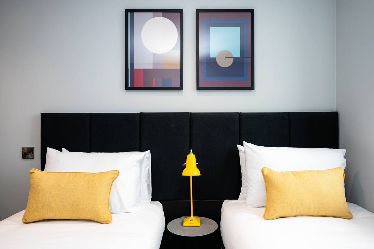 A hotel room with a black headboard, two white pillows, a yellow lamp, and two framed artworks on the wall.