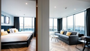 Una habitación de hotel moderna con una cama grande, un sofá, una mesita y vistas a la ciudad a través de amplios ventanales.