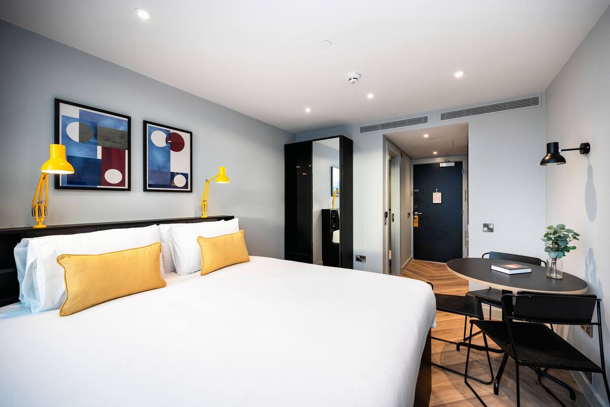 A modern hotel room with a large bed, bedside lamps, a desk with a chair, and artwork on the wall.