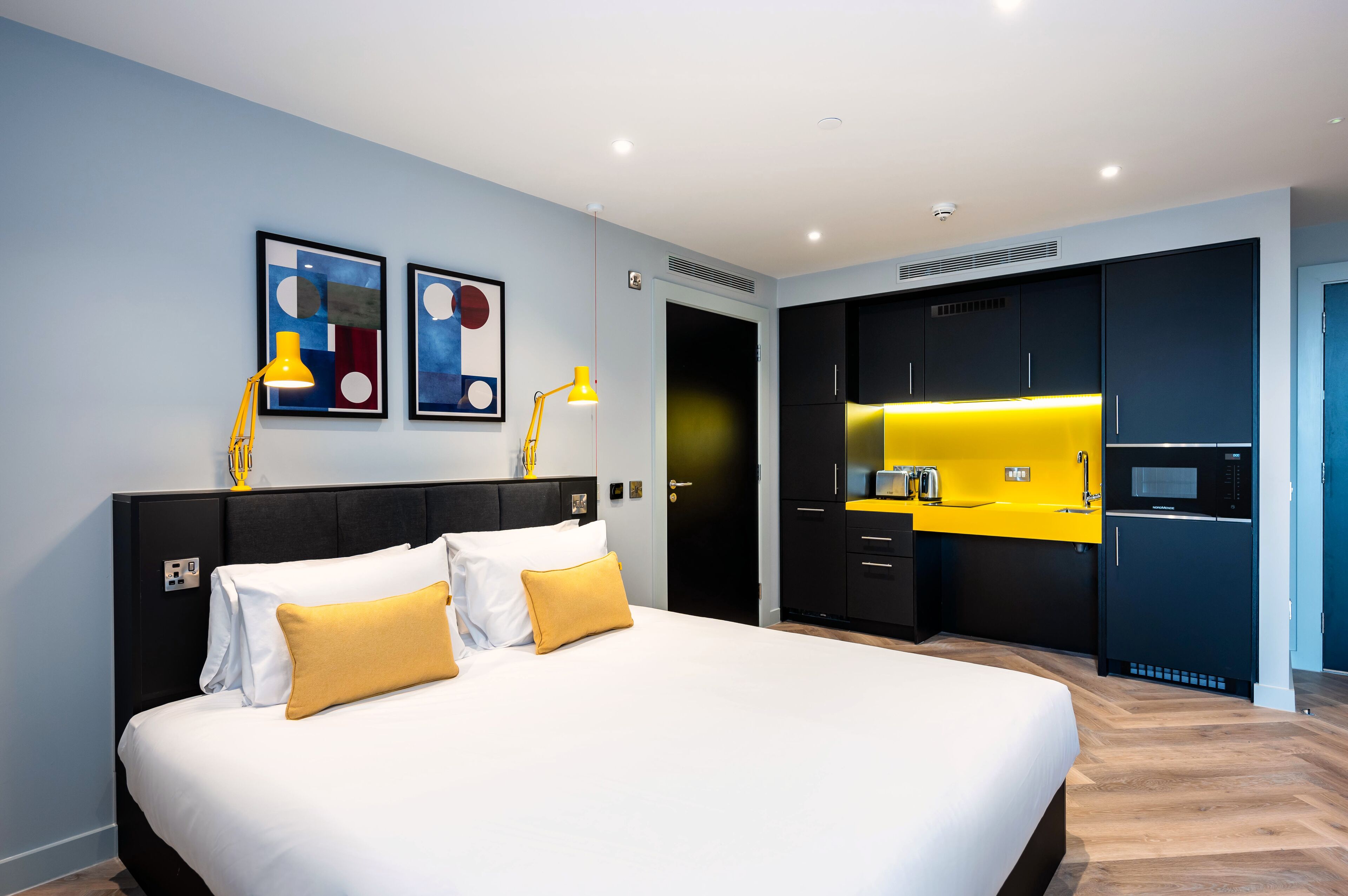 A modern hotel room with a large bed, a small kitchen area, and artwork on the walls.