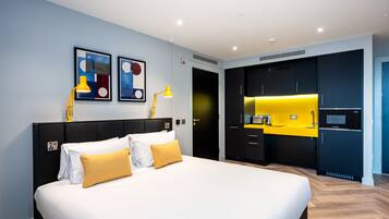 A modern hotel room with a large bed, a small kitchen area, and artwork on the walls.