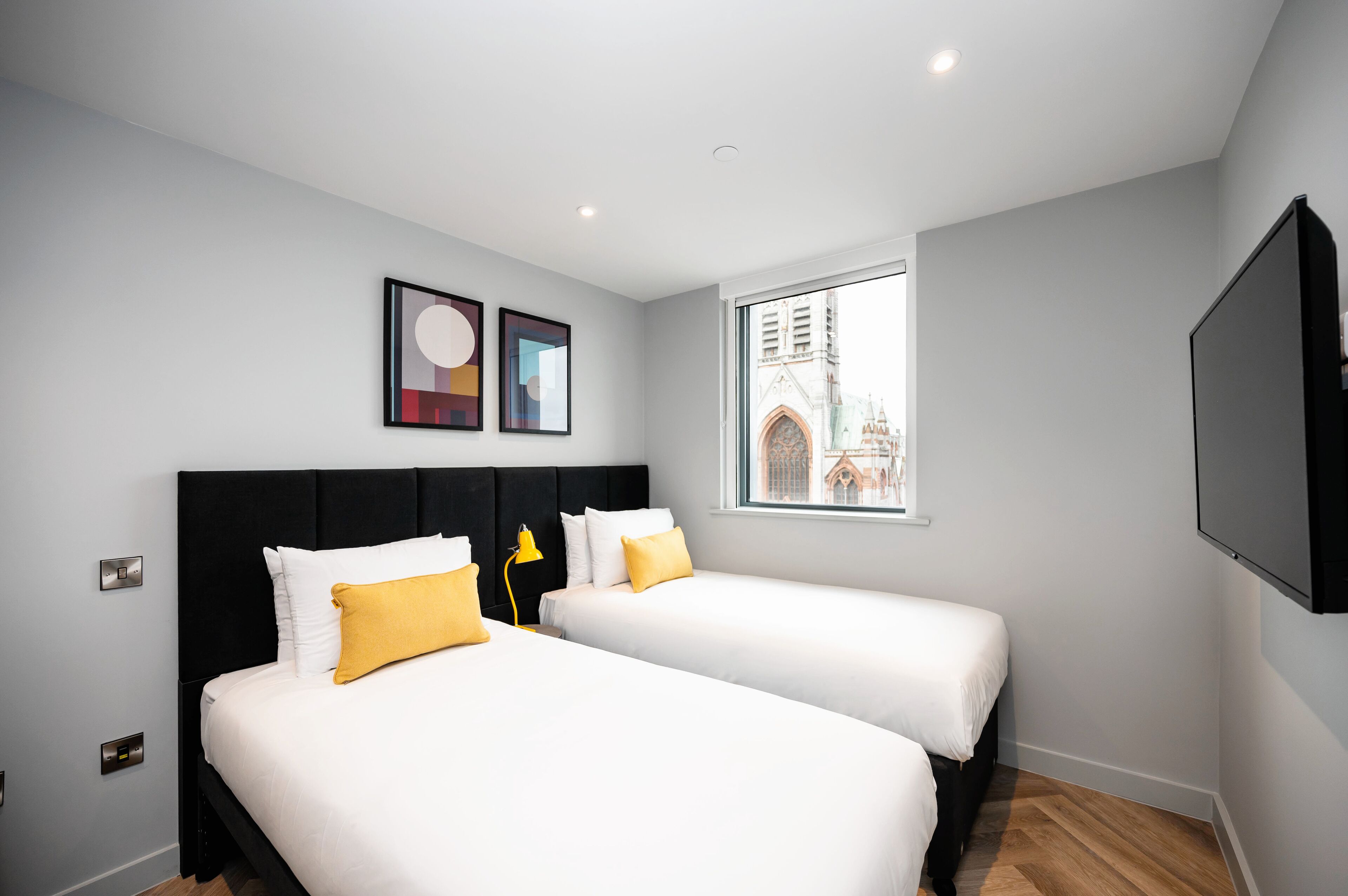 Habitación de hotel con dos camas, un ventanal con vista a un edificio y un televisor de pantalla plana montado en la pared.