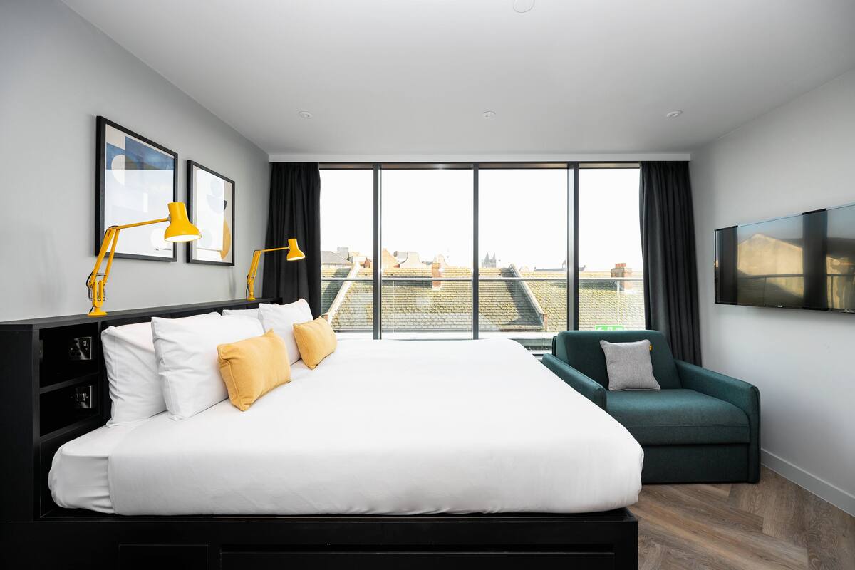A modern hotel room with a large bed, a green armchair, a flat-screen TV, and a city view through large windows.