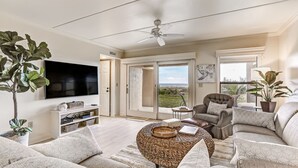 Ein geräumiges Wohnzimmer mit Flachbildfernseher, bequemen Sitzgelegenheiten, Deckenventilator und großen Fenstern mit Blick ins Freie.