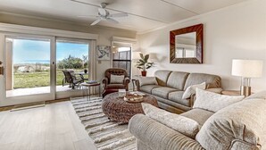 Ein geräumiges Wohnzimmer mit einer großen Couch, einem Couchtisch und einem Deckenventilator. Eine Glastür öffnet sich zu einem Patio mit Gartenmöbeln.