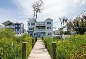 A wooden pathway leading to a multi-story residential building with balconies and a view of the surrounding greenery. - Pier, Pool, Sunsets • Luxe Soundfront Retreat (Corolla)
