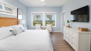 A bedroom with a large bed, a desk, a chair, a TV, and a window with a view of the outdoors. - Pier, Pool, Sunsets • Luxe Soundfront Retreat (Corolla)
