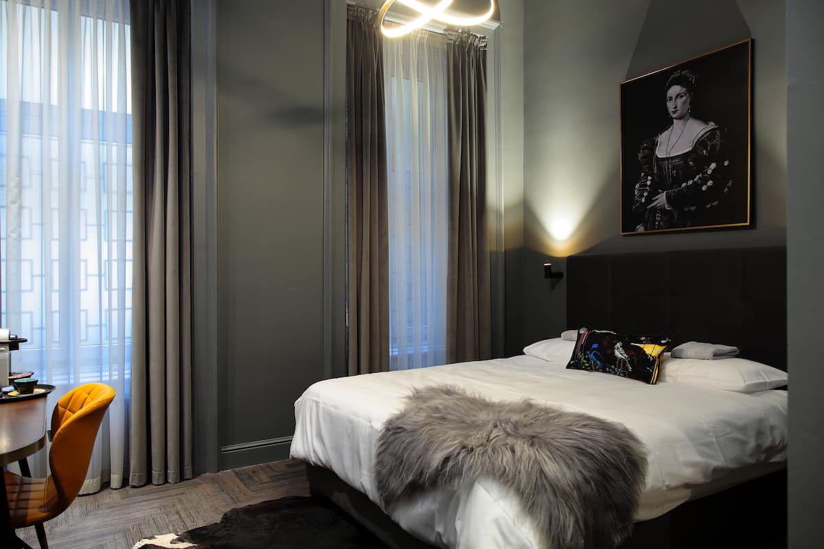 A modern hotel room with a large bed, a desk with a chair, and a painting on the wall.