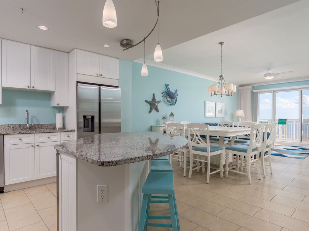 A kitchen with white cabinets, a granite countertop, and a stainless steel refrigerator. There is a dining area with white chairs and a table.