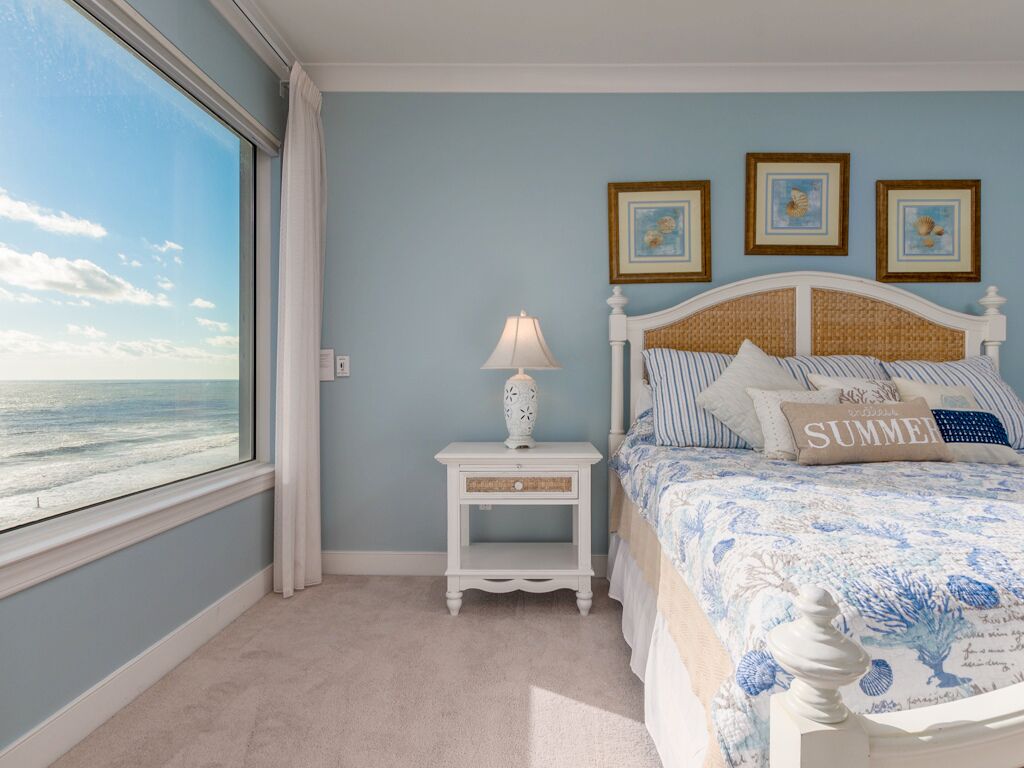 A bedroom with a large window, a bed with a floral bedspread, a nightstand with a lamp, and three framed pictures on the wall.