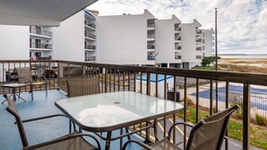 Uma varanda com mesa e cadeiras de vidro, com vista para um parque de estacionamento e um edifício com várias varandas.