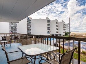 A balcony with a glass table and chairs, overlooking a parking area and a building with multiple balconies.