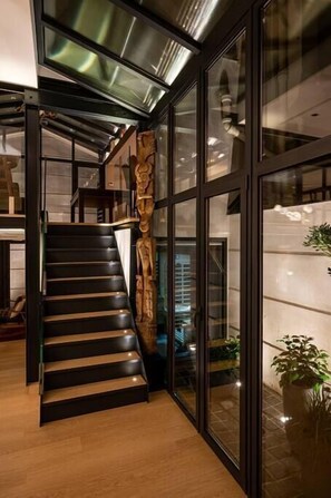 Un escalier moderne avec des rampes en verre et une main courante en bois, menant à un espace situé au deuxième étage et offrant une vue sur l’intérieur.