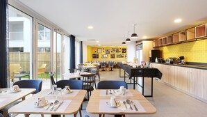 Un comedor moderno con mesas dispuestas para el desayuno, una zona de cocina con armarios de madera y una pared de ladrillo amarillo con obras de arte.