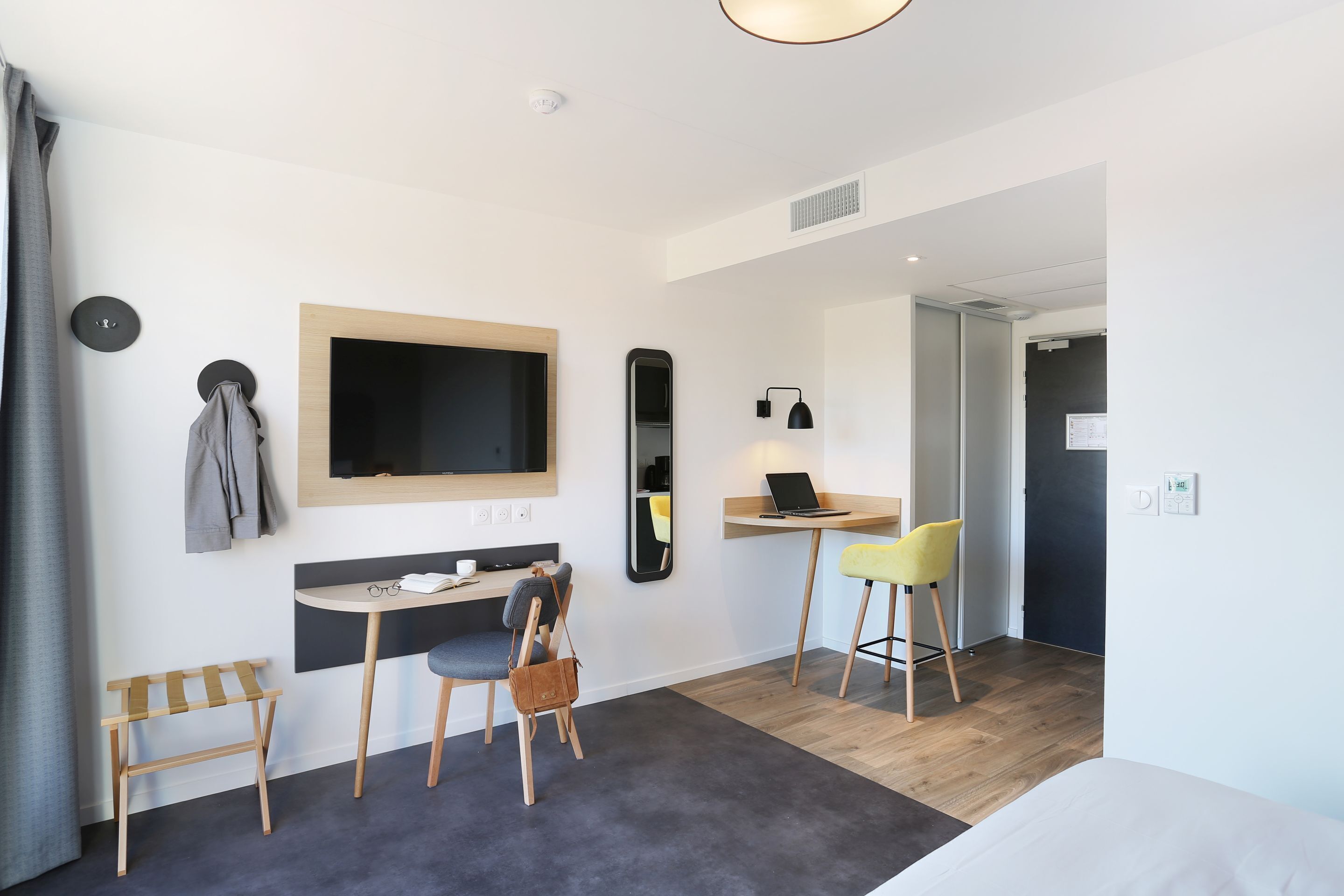 Een moderne hotelkamer met een bed, een bureau met een laptop, een stoel, een klein tafeltje met glazen, een kapstok en een spiegel.