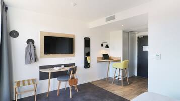 Een moderne hotelkamer met een bed, een bureau met een laptop, een stoel, een klein tafeltje met glazen, een kapstok en een spiegel.