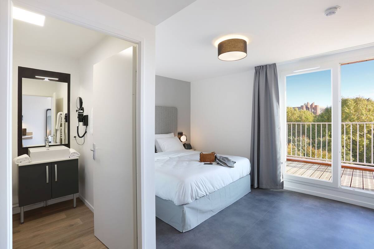 A modern hotel room with a large bed, a bathroom with a sink and mirror, and a balcony with a view.