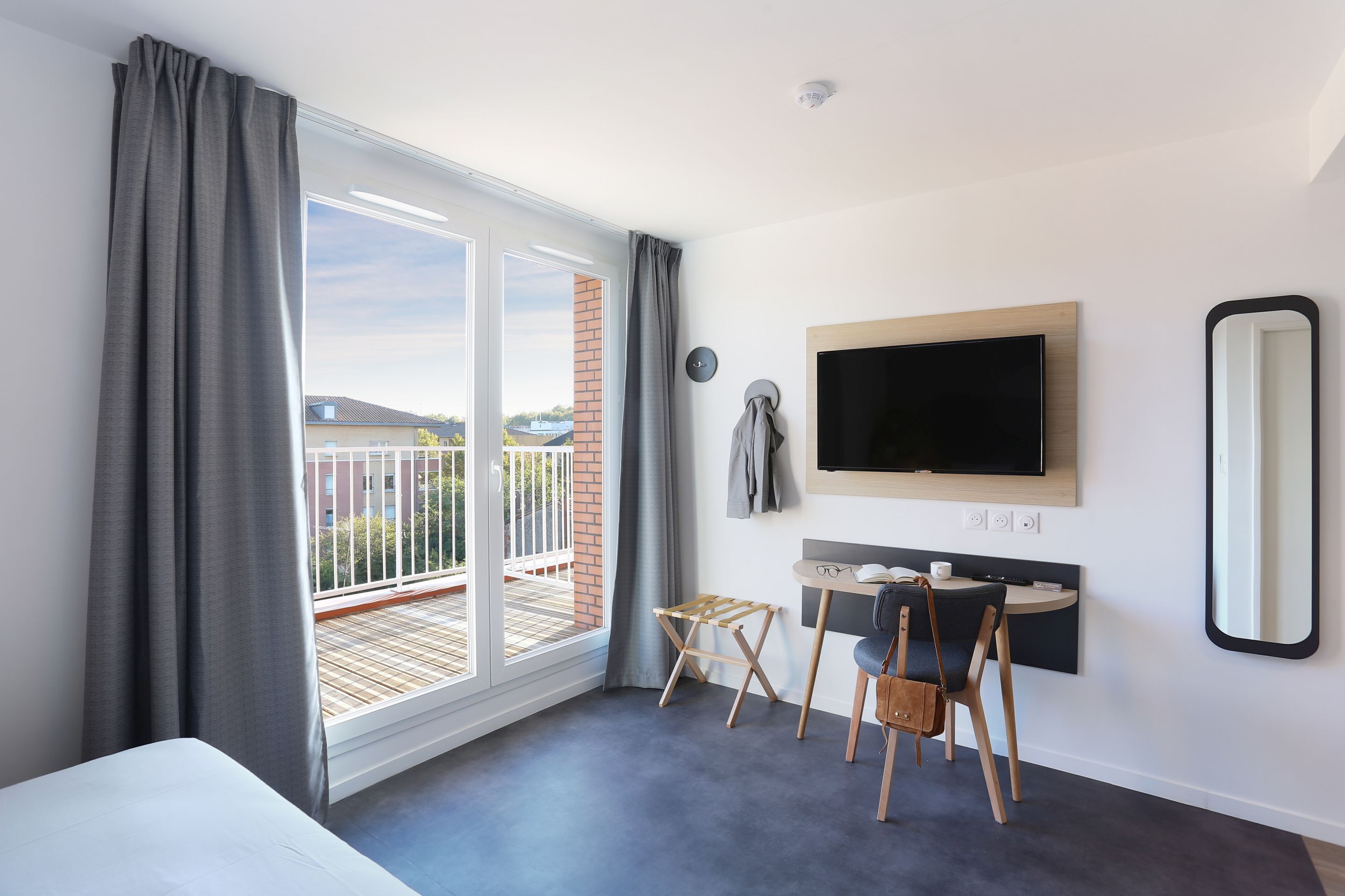 Een moderne hotelkamer met een grote schuifglazen deur naar een balkon, een flatscreen televisie aan de muur, een klein rond tafeltje met een stoel en een compact badkamergedeelte.