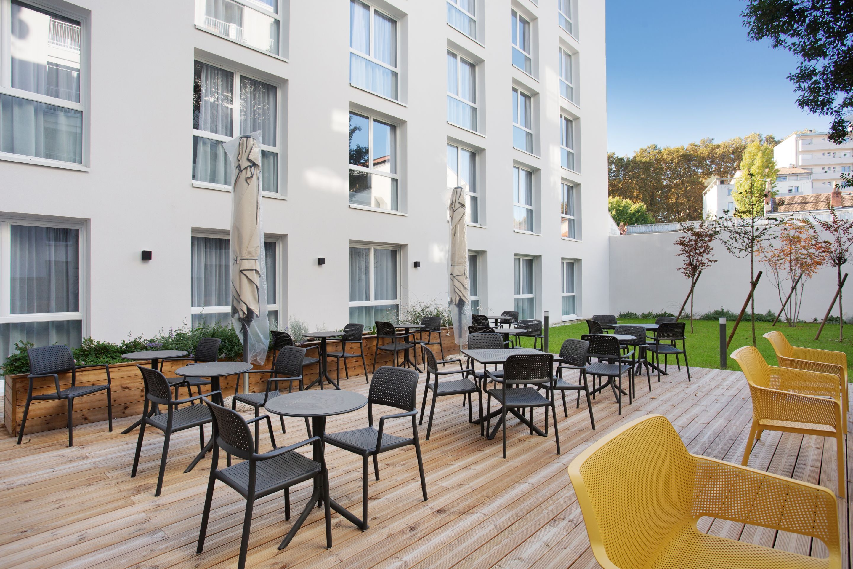 Une terrasse extérieure avec des tables et des chaises, une plateforme en bois et un bâtiment blanc doté de nombreuses fenêtres.