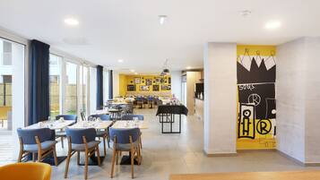 A modern restaurant with a bright yellow and black mural, wooden tables and chairs, and large windows allowing natural light.