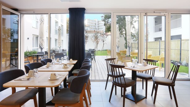 Un comedor con mesas y sillas, ventanas amplias y vistas a un edificio y follaje exterior.