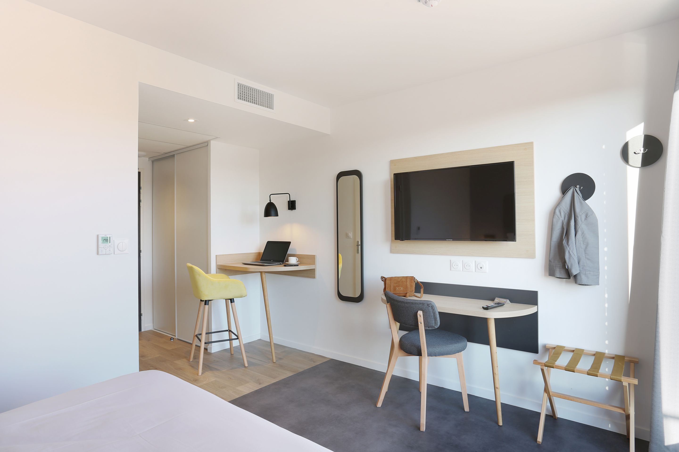 Ein modernes Hotelzimmer mit einem Bett, einem Schreibtisch mit einem Laptop, einem Spiegel und einem Fernseher.