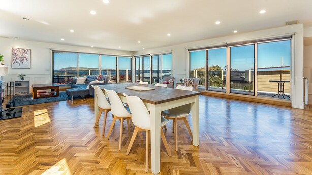 Een ruime woonkamer met een houten visgraatvloer, een grote eettafel met stoelen, een bank en een uitzicht door grote ramen.