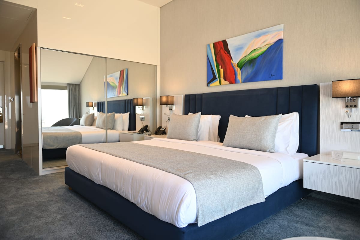 A modern hotel room with a large bed, a bedside table, a mirror, and a painting on the wall.