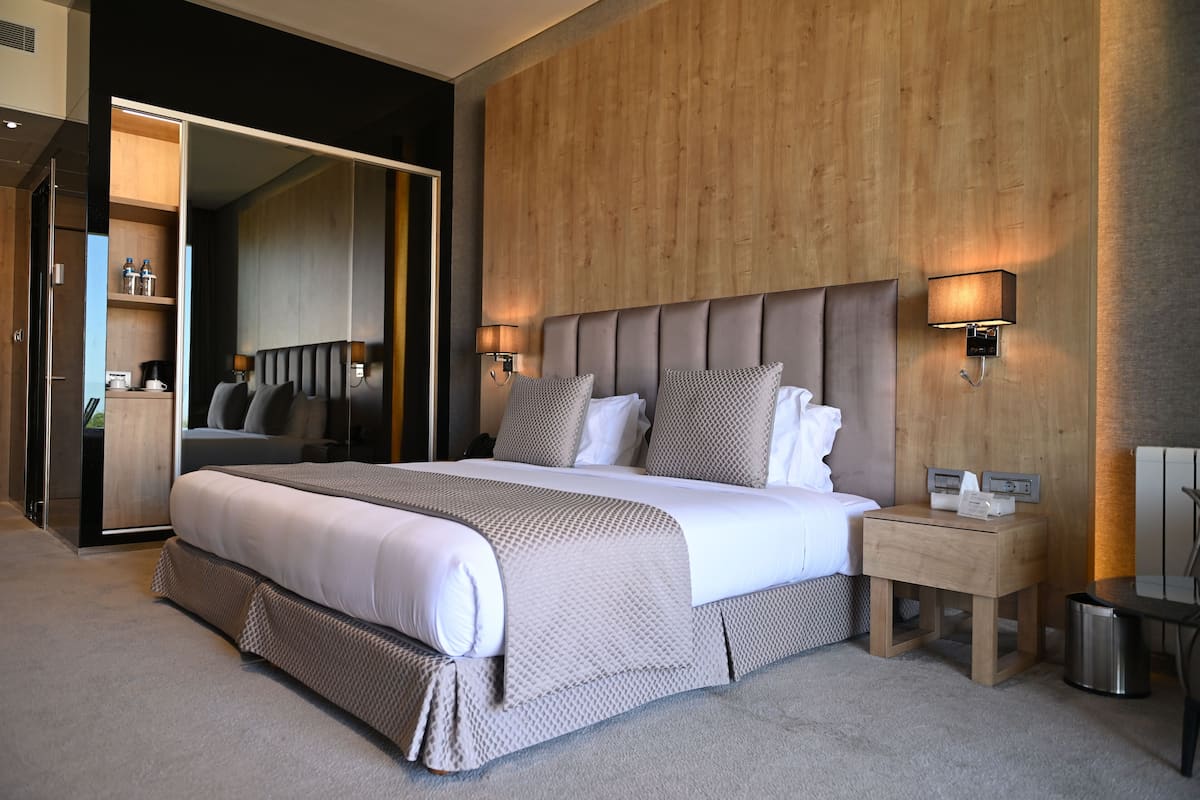 A modern hotel room with a large bed, wooden panel walls, and a small bedside table with a lamp.