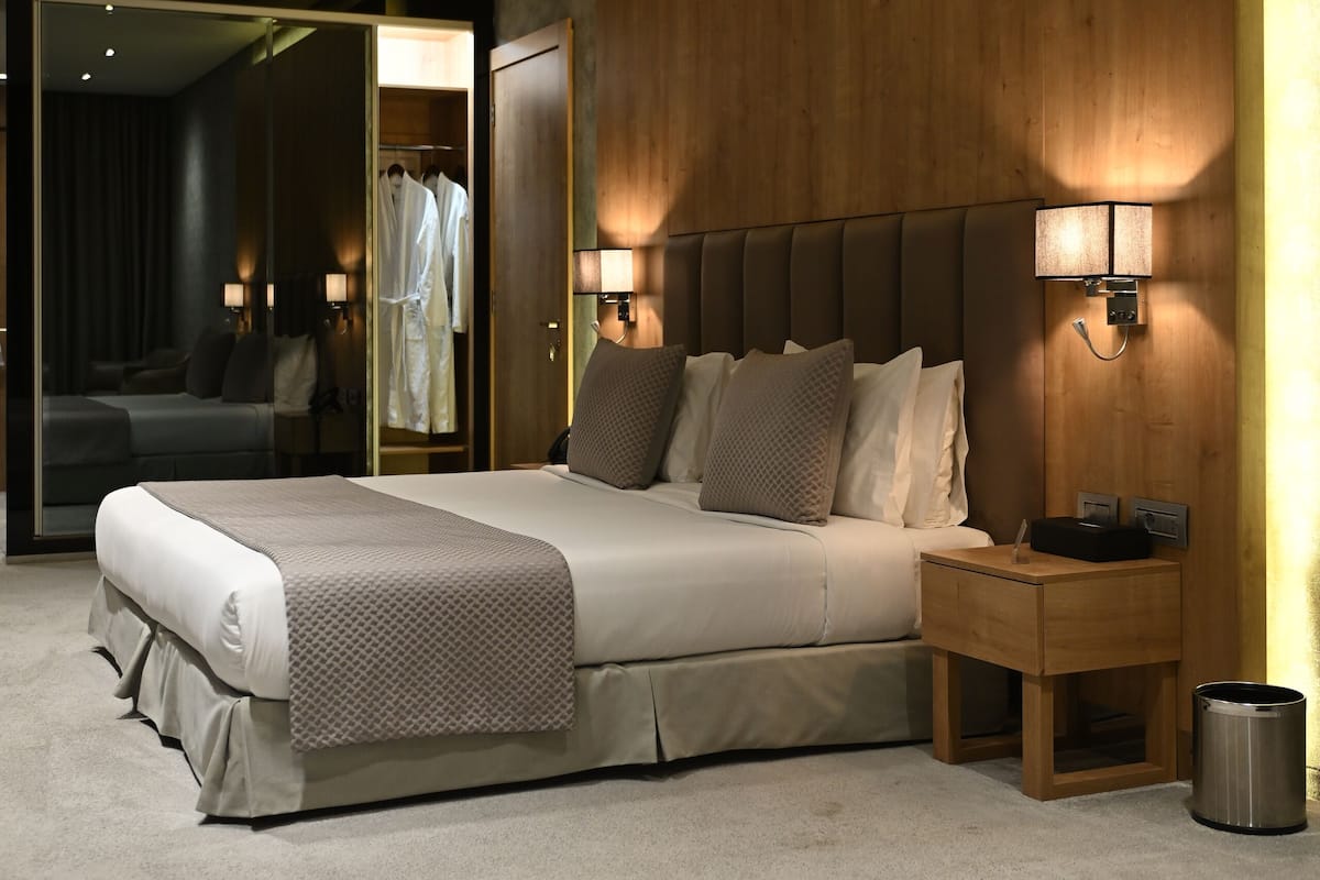 A modern hotel room with a large bed, bedside lamps, a wooden headboard, a nightstand, and a mirror.