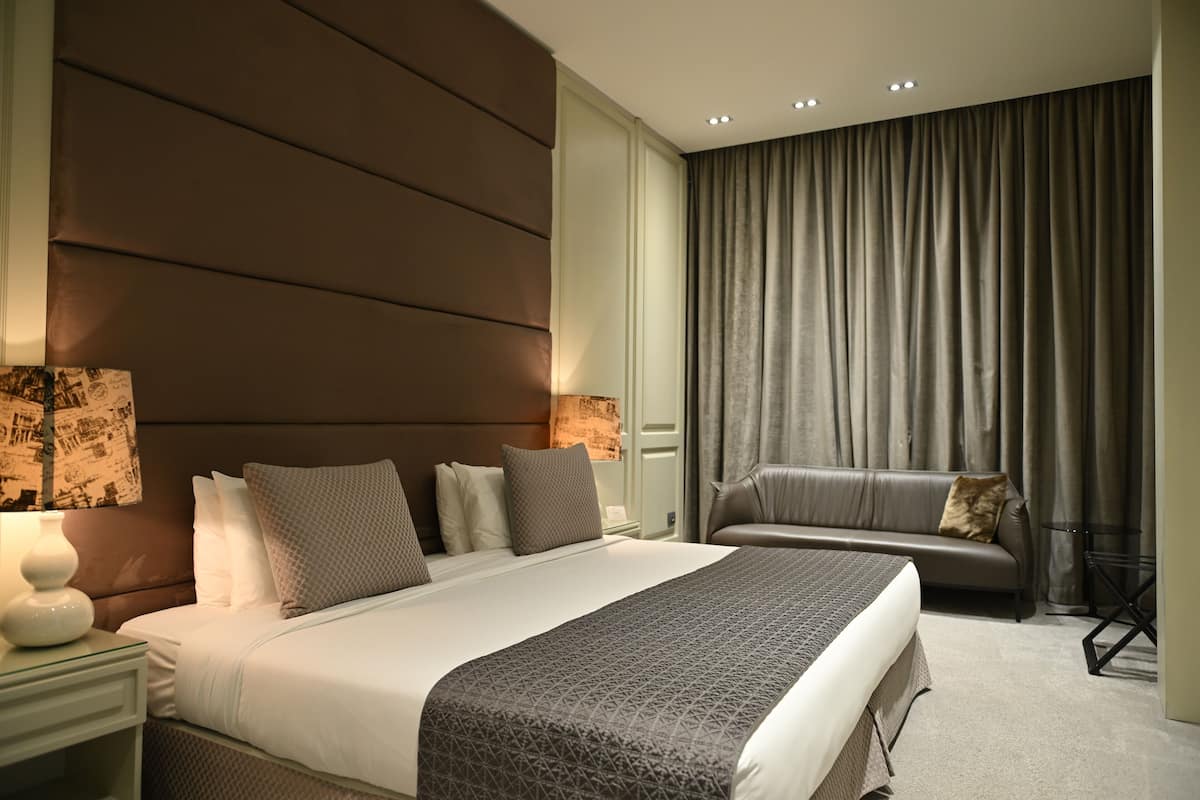 A modern hotel room with a large bed, a sofa, and a nightstand with a lamp.
