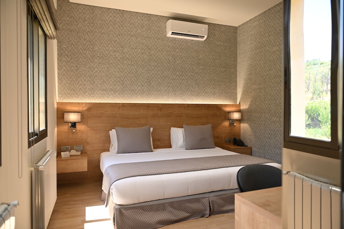A hotel room with a large bed, bedside lamps, a wooden headboard, and a view of greenery outside.