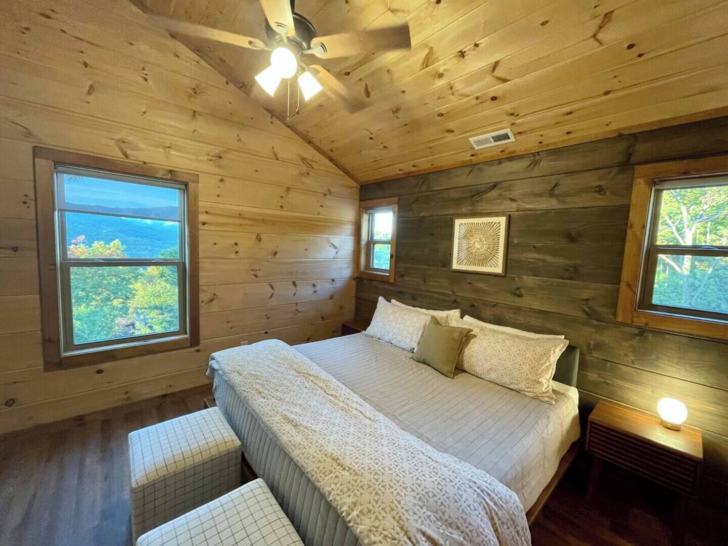 Ein Schlafzimmer einer Holzhütte mit Bett, zwei Fenstern, Deckenventilator und Nachttisch mit Lampe.