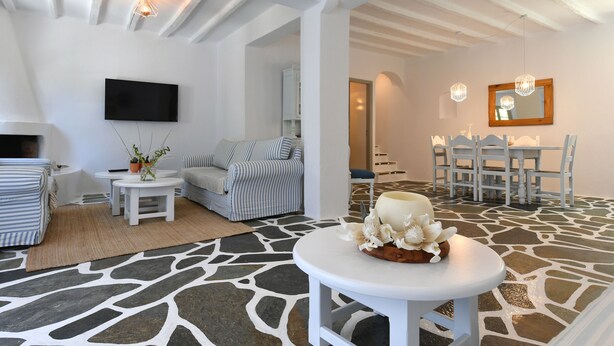 A modern living room with a stone-patterned floor, a white sofa, a television, and a dining area with white furniture. - Santa Flora 3 bedroom House (Paros)