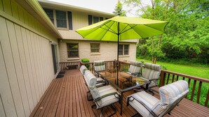A wooden deck with a glass-top table and six striped chairs, a green umbrella, and a stone building in the background.