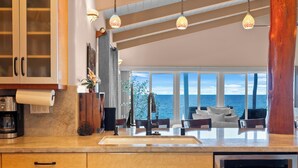 A modern kitchen with a large window overlooking the sea, a dining area with a view, and a wooden cabinet with a coffee machine.