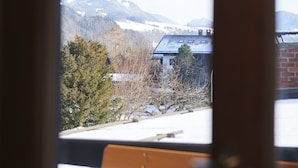Una vista attraverso una finestra con cornice in legno, con un edificio a tetto spiovente, neve a terra e un paesaggio montuoso sullo sfondo.