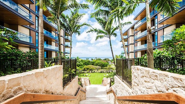 A hotel with a stone staircase leading to a pool area, surrounded by palm trees and a view of the ocean.
