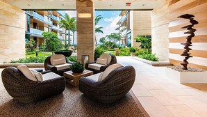 A modern lobby with wicker furniture, a central pillar, and a view of a landscaped outdoor area with buildings in the background.
