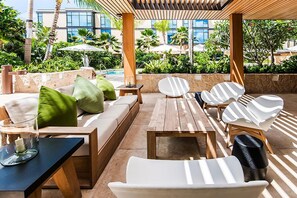 Een zonnig buitenterras met een zwembad, houten tafels en lounge fauteuils.