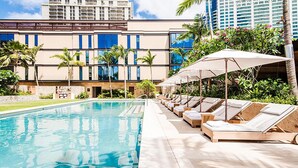A swimming pool with lounge chairs and umbrellas, surrounded by palm trees and a building with multiple floors.