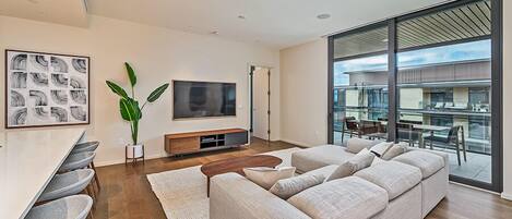 A modern living room with a large sofa, a dining area with chairs, a TV, and a large window offering a view of the building's exterior.